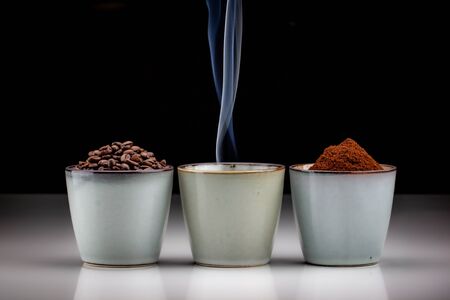 3 rustic coffee cups with coffee grounds, smoking coffee, and ground coffee. Standing on a white reflective table fading into a black backgroundの写真素材