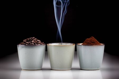 3 rustic coffee cups with coffee grounds, smoking coffee, and ground coffee. Standing on a white reflective table fading into a black backgroundの写真素材