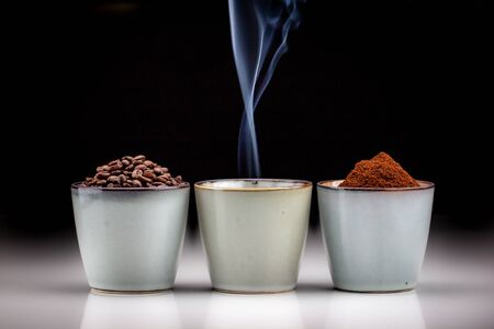 3 rustic coffee cups with coffee grounds, smoking coffee, and ground coffee. Standing on a white reflective table fading into a black backgroundの写真素材