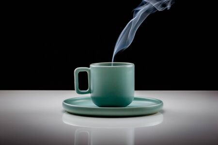 Pastel green colored coffee or tea cup with hot liquid, smoke and steam, black background. Cup standing on a slightly reflective table that fades into a black backgroundの写真素材