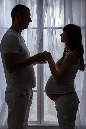 Young couple waiting for a baby face off bellies, standing at the windowの写真素材