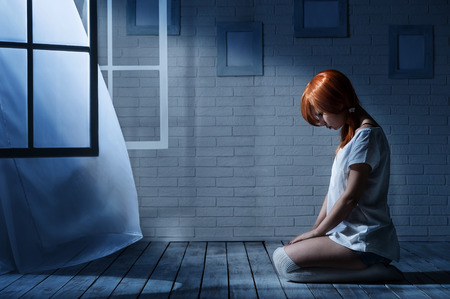Lonely girl sits in an empty dark room opposite the windowの写真素材