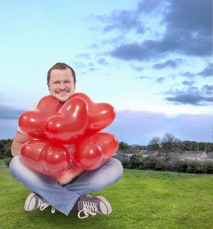 Young man sitting with red balloons with green lawn field in backgroundの写真素材