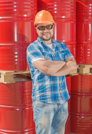 Worker in safety helmet and sunglasses standig in front of metal barrelsの写真素材