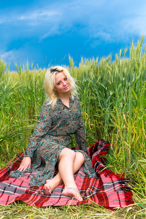 Attractive young blonde woman sitting on plaid in rye fieldの写真素材