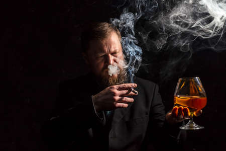 Solid confident bearded man in suit with glass of whisky and cigar with fumeの写真素材