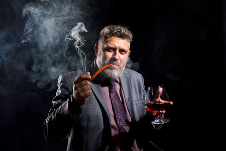 Solid confident bearded man in suit with glass of whisky smoking pipeの写真素材