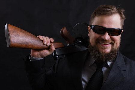 Mature bearded man in sunglasses dressed in suit with tommy gun