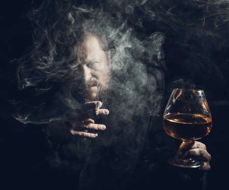 Solid confident bearded man in suit with glass of whisky and cigar with fumeの写真素材