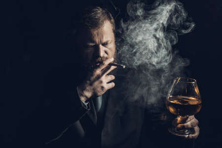 Solid confident bearded man in suit with glass of whisky and cigar with fumeの写真素材