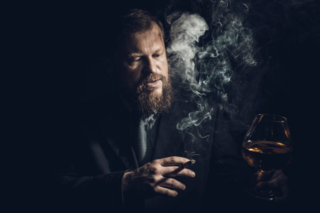 Solid confident bearded man in suit with glass of whisky and cigar with fumeの写真素材