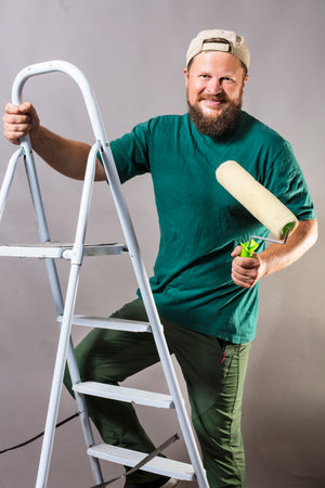 Joyful bearded foreman in green t-shirt with brush roller and ladderの写真素材