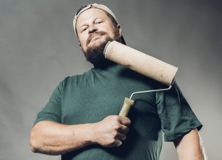 Joyful bearded craftsman in green t-shirt having fun with paint rollerの写真素材