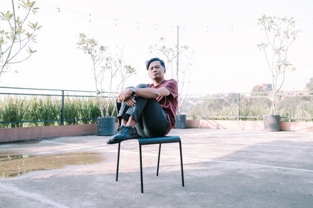 A Southeast Asian man sits on a chair on a building balcony, enjoying the view or relaxing. Perfect for themes of leisure, urban living, and outdoor spaces.の写真素材