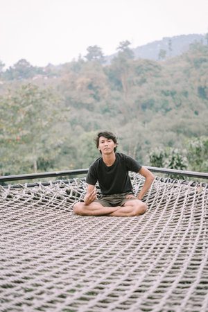 A Southeast Asian man relaxing on a net, with a scenic view below him, enjoying a peaceful and...