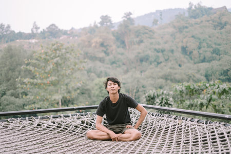 A Southeast Asian man relaxing on a net, with a scenic view below him, enjoying a peaceful and serene moment amidst natureの写真素材
