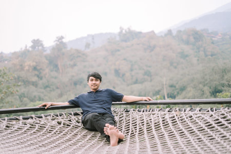 A Southeast Asian man relaxing on a hammock-style net with a breathtaking view in the background,...