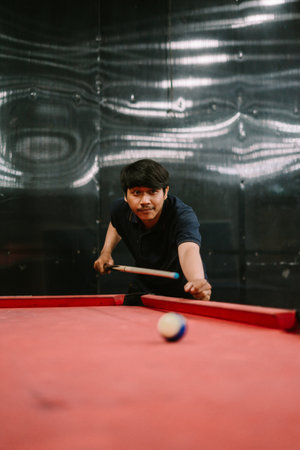 A Southeast Asian man playing billiards, focused on aiming the cue ball during a game, captured in a relaxed and engaging setting.の写真素材