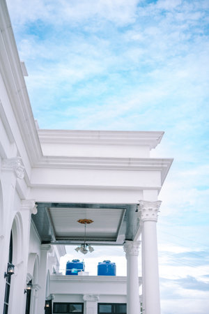 A luxurious white colonial-style building surrounded by vibrant greenery and a landscaped lawn, with a clear pathway leading to its entrance under a bright, cloudy skyの写真素材