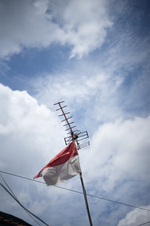 Indonesian flag waving beneath a television antenna against a cloudy sky.
