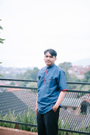 A portrait of an Indonesian man standing outside wearing traditional clothing, thoughtfully looking...