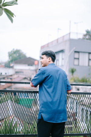 A young man gazes out at the city view from a rooftop balcony, caught in a moment of thoughtful contemplation. The urban setting provides a backdrop for this scene of modern lifestyle. This image conveys a sense of peace, solitude, and introspection, perfect for evoking feelings of inspiration and positive thinking.の写真素材