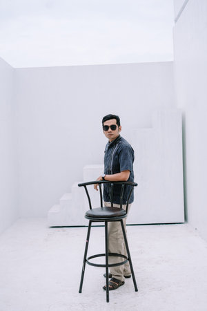 A stylish man wearing sunglasses leans casually on a chair in a minimalist white studio setting. The...
