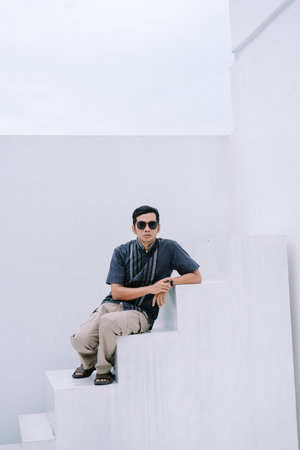 A stylish man wearing sunglasses poses thoughtfully on a white block construction outdoors. The minimalist architecture and urban setting add to the trendy and contemporary feel of the image. The daylight emphasizes the details of the scene.の写真素材