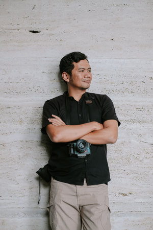 A thoughtful photographer leans against a textured stone wall, camera hung around his neck. With arms crossed, he gazes thoughtfully, seemingly contemplating his next shot, bathed in the soft outdoor light. He exudes confidence and expertise.の写真素材
