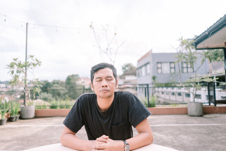 A serene Asian man sits with closed eyes, practicing mindfulness and meditation outdoors. He seeks inner peace and a calm, balanced state amidst the urban landscape. Rooftop setting provides a quiet escape for thoughtful reflection and personal wellbeing.の写真素材