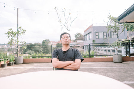 A man sits pensively with crossed arms on an urban rooftop terrace. Overlooking the city, he exudes a confident and thoughtful demeanor, blending casual style with a touch of seriousness.の写真素材