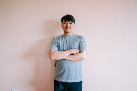 Portrait of a young asian man with a mustache, arms crossed, looking confidently at the camera. He is wearing a grey t-shirt against a neutral backdrop. He shows a positive attitude.の写真素材