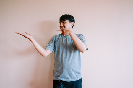 Young man presenting or showing something with his hand. Smiling Asian guy in casual wear gesturing to empty space with a finger pointing. Advertisement and promotion concept, with plenty of copy space.の写真素材