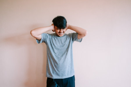 A young man experiencing discomfort covers his ears in an attempt to block out an irritating or loud noise. He is visibly stressed and upset by the sound, expressing frustration and pain.の写真素材