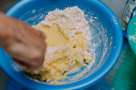 Close-up image shows hand mixing flour and batter in a blue bowl to prepare baking ingredients. This...
