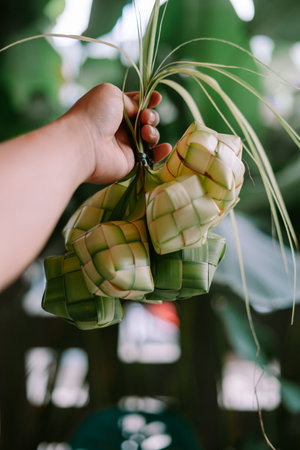 A hand holds a bunch of ketupat, a traditional rice dumpling served during Eid al-Fitr. This dish is commonly found in Southeast Asia, especially Indonesia, Malaysia, Brunei, and Singapore. Ketupat is made from rice packed inside a woven palm leaf pouch, then boiled. It is often served with meat dishes.の写真素材