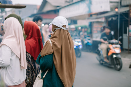 A glimpse into the daily life of a Southeast Asian city. A group of women wearing hijabs walks down a busy street filled with motorcycles. The scene depicts cultural diversity and a bustling urban environment.の写真素材