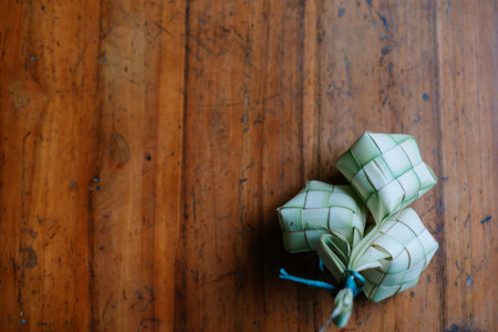 Three handwoven ketupat, a rice dumpling, lie on a rustic wooden table. A traditional Southeast Asian delicacy for festive occasions, these woven palm leaf pouches are filled with fragrant rice and boiled to perfection.の写真素材