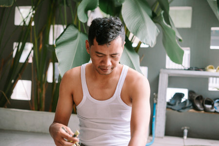 An asian man in a tank top concentrates on trimming plants outdoors. Focused on the task, he tends to his garden in a relaxed, tropical environment. Represents a hobby, wellbeing, and simple domestic life.の写真素材