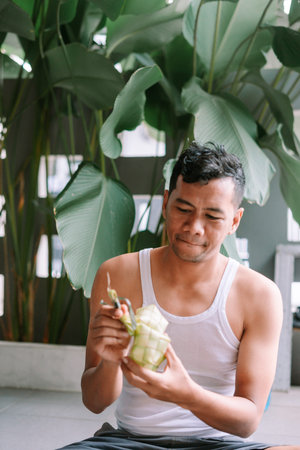 A man carefully trims a freshly cooked Ketupat. Ketupat is a rice dumpling packed inside a...