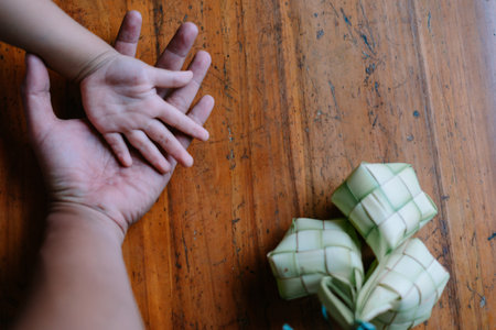 Image captures a child's hand resting on a parent's hand, symbolizing the bond during Eid al-Fitr celebration with Ketupat. It represents themes of family, tradition, cultural heritage, and the spirit of togetherness during festive times.の写真素材