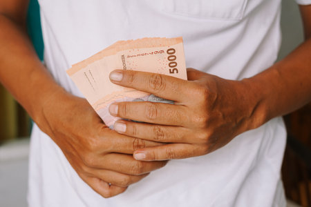 Close-up of a man holding a stack of 5000 Indonesian Rupiah banknotes. This image conveys financial concepts such as wealth, savings, payments, and investment in Indonesian currency.の写真素材