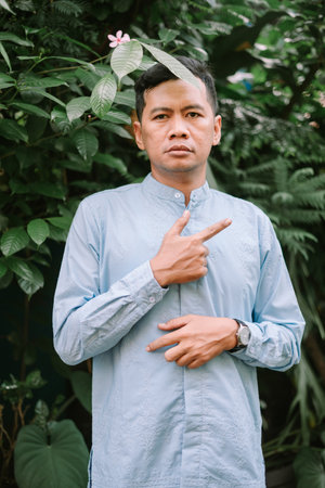 An Asian man in a light blue shirt stands outdoors, pointing to the side. Surrounded by lush greenery, his serene expression and focused gaze create a captivating portrait. Perfect for illustrating guidance, presentation, or natural wellness concepts.の写真素材