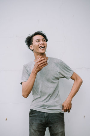 A happy young man wearing a gray shirt smiles broadly against a textured white wall with a joyful...
