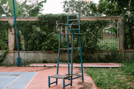 Weathered volleyball referee chair stands alone on an outdoor court, set against a lush green backdrop of plants.の写真素材