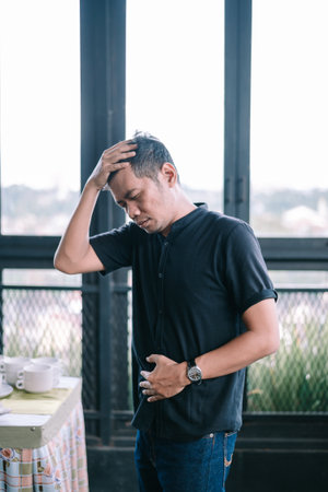 Man experiencing stomachache and headache in bright indoor setting, looks unwell and concerned. He is holding his stomach and head in distress.の写真素材