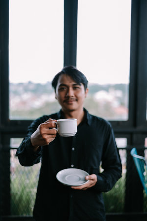 Man enjoys his beverage. Stylish asian male holding a coffee cup and saucer, smiling in front of the windowの写真素材