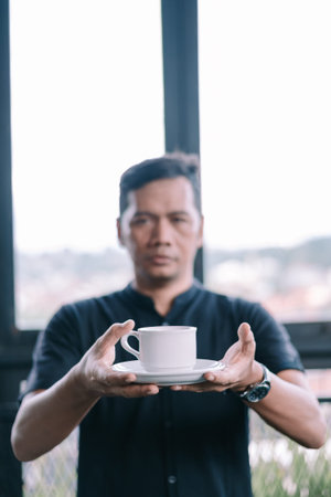 A man presents a cup of coffee, selective focus. Perfect for illustrating a coffee break or hospitality with its blurred modern settingの写真素材