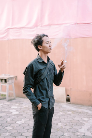 Young man smoking a cigarette outdoors. He is standing in a contemplative poseの写真素材