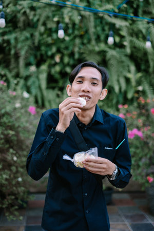A cheerful man enjoys a snack outdoors amidst lush greenery. He is smiling and happy to be enjoying traditional indonesian street foodの写真素材